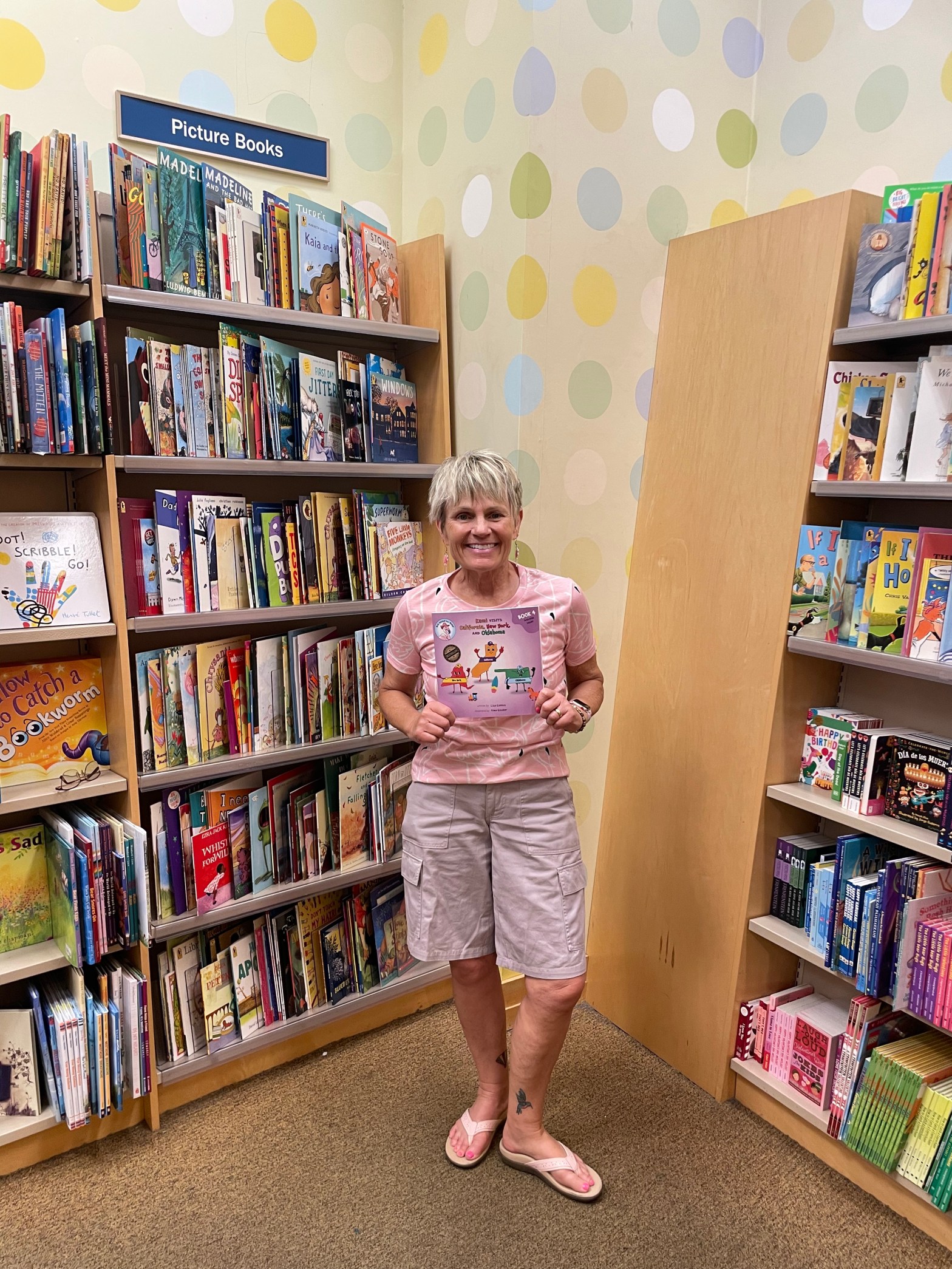 Author Lisa Galdos smiling in a bookstore children’s section, holding her BIG HUGS for Kids book while standing between shelves of picture books.