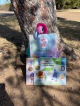 Children’s books and an activity book displayed outdoors against a tree with a pink flamingo plush, promoting Kami’s Collection.