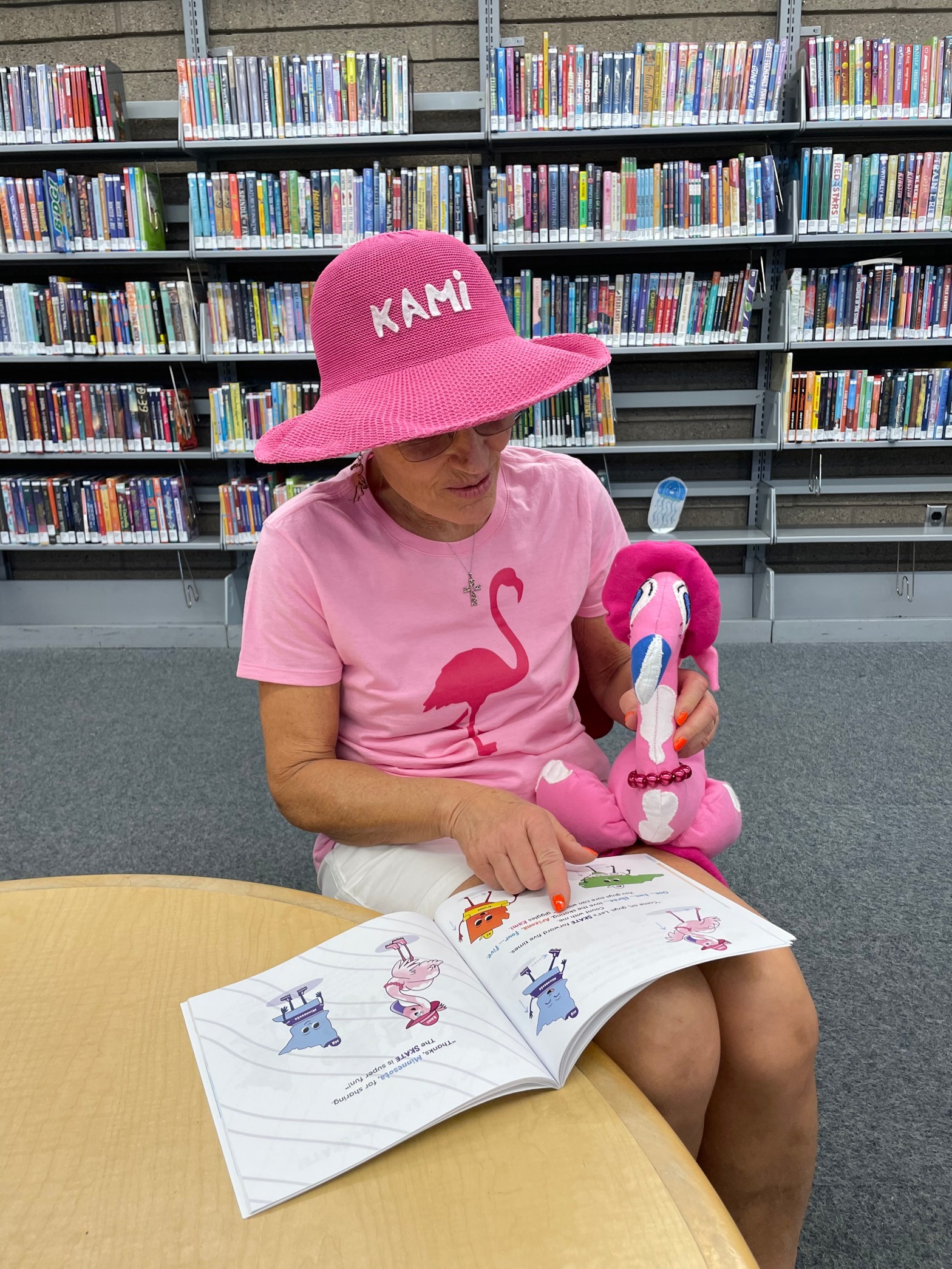 Woman wearing a pink hat and shirt with a flamingo design reading a children’s book while holding a flamingo plush in a library.